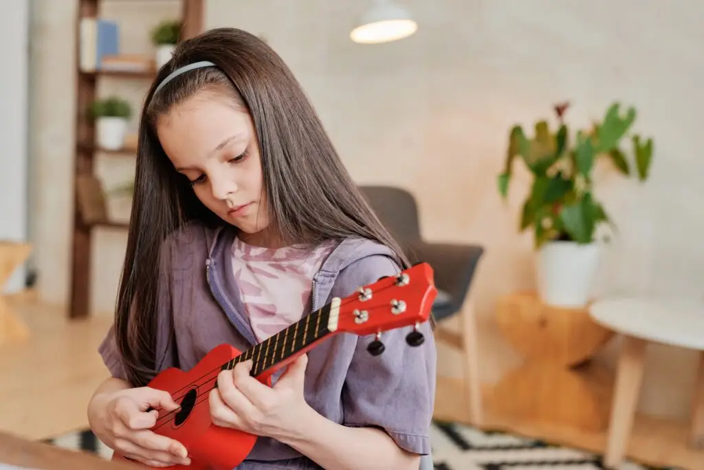 Young girl learning to play a red ukulele indoors, focusing on the instrument.