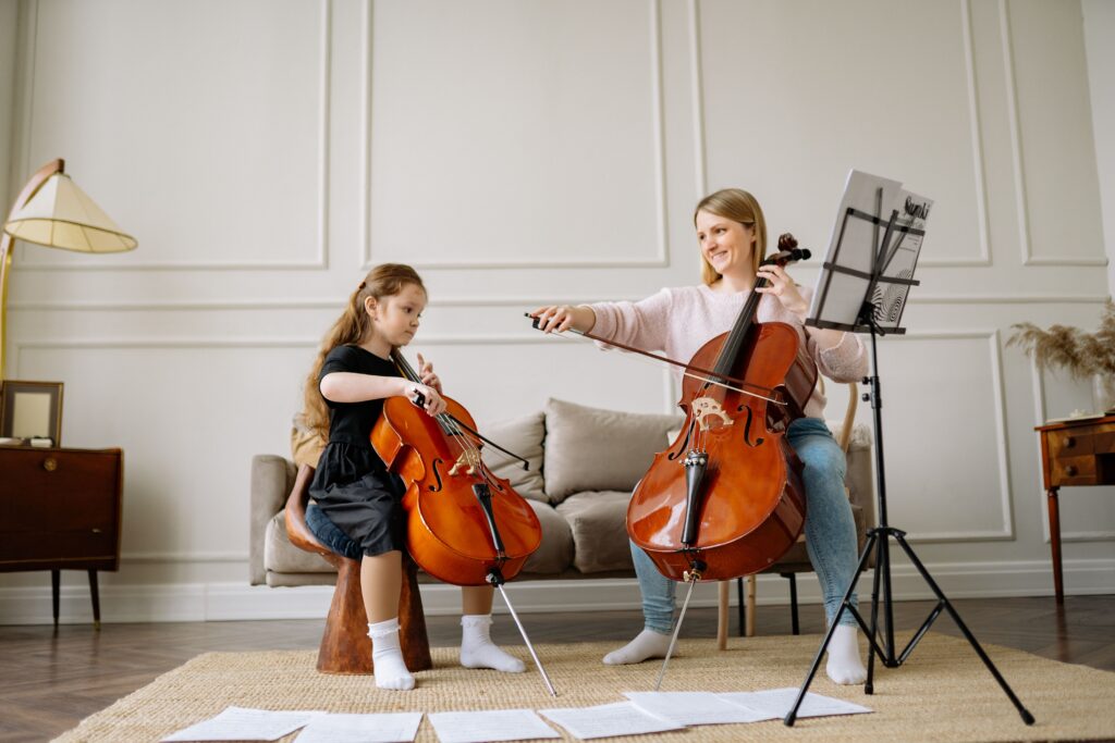 A woman and a girl playing cellos together in a cozy living room setting, fostering music education and bonding.
